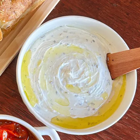 Tigela de coalhada seca cremosa, com fio de azeite e espátula de madeira, ao lado da focaccia