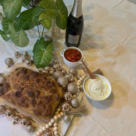 Focaccia dourada em tábua ao lado de coalhada seca, tomate confit e uma garrafa de espumante, em mesa decorada para o Réveillon